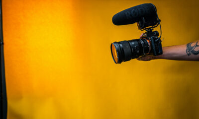 A man with a tattoo holding out a black DSLR camera in front of an orange background.