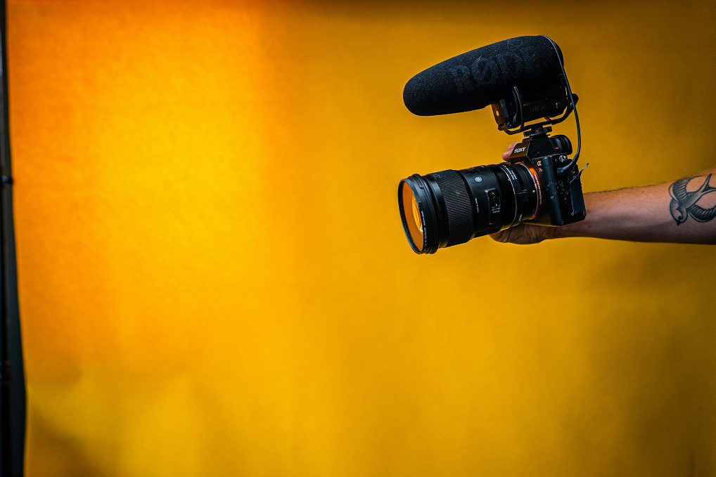 A man with a tattoo holding out a black DSLR camera in front of an orange background.